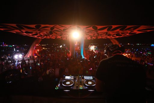 A DJ performs for a large crowd at night under a patterned canopy at Burning Man 2019. The DJ is silhouetted against the bright lights of th