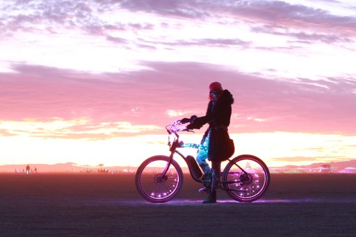 A person stands with a bicycle adorned with purple and blue lights against a dramatic pink and orange sunset sky. The scene was captured on