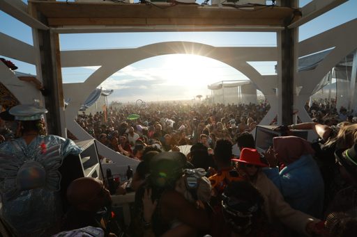 A large crowd gathers outdoors at sunset, viewed from a raised platform with white architectural elements. The sun flares brightly in the sk