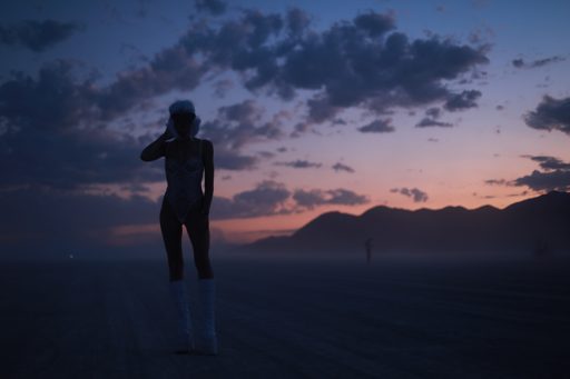 A silhouette of a woman in a leotard and tall boots stands in a vast, dusty landscape at dusk. She wears a white furry hat and earmuffs, and