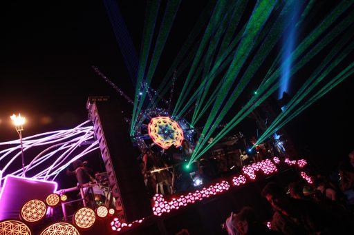 Green and blue lasers illuminate a stage setup with a large geometric art installation during a night event in 2016. The scene features a cr