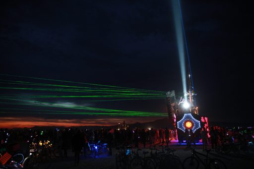 A large art installation with a powerful beam of light shooting upwards into the night sky is illuminated by lasers and colorful lights, sur