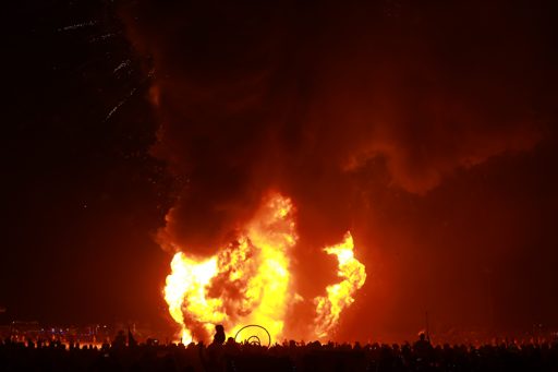A massive explosion of fire and smoke erupts from the Man structure at Burning Man 2019, witnessed by a large crowd silhouetted against the