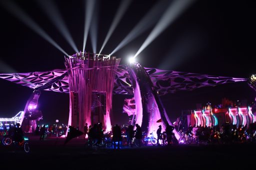 The "Temple" structure at Burning Man 2019 is illuminated with purple lights and spotlights from above, with a crowd of people and bicycles