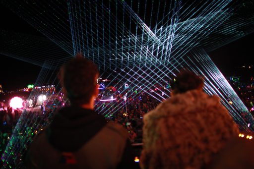Two people stand with their backs to the camera, looking out at a large crowd illuminated by a complex laser light show at night. The scene