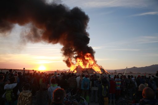 A large wooden effigy burns intensely against a sunset sky, drawing a crowd of onlookers at the Burning Man festival in Black Rock City, Nev