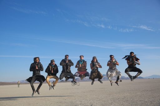 Seven individuals in costumes jump in unison with their hands pressed together in a prayer-like gesture on the dry lakebed of Black Rock Des