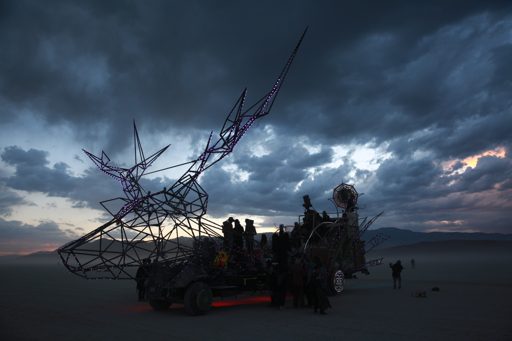 A large metal sculpture resembling a deer's head, adorned with purple lights, is mounted on a vehicle in the Black Rock Desert at dusk. The