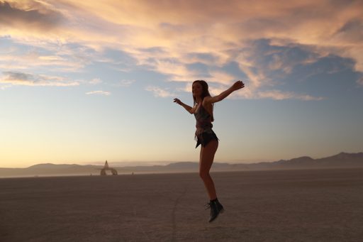 A woman jumps in the air in front of the Temple structure at sunset during the Burning Man festival. The sky is filled with dramatic clouds