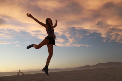 A woman in a patterned top and black skirt jumps in the air against a dramatic sunset sky with wispy clouds. In the distance, a triangular s
