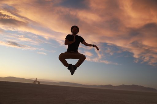 A man in silhouette jumps in the air with his hands in a prayer-like pose against a dramatic sunset sky during the Burning Man 2019 festival