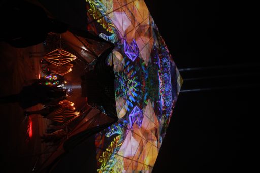 A kaleidoscopic projection of colorful geometric patterns and floral motifs is cast onto a mirrored surface, reflecting a hand holding a str