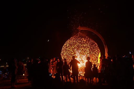 A crowd gathers at night to watch a large, geodesic sphere sculpture ignite, sending sparks into the dark sky. A crescent moon is visible ab