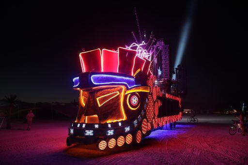 A large, illuminated art car resembling a train, adorned with neon lights and speakers, is parked on the playa at night during the 2017 Burn