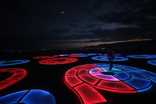 A person stands on a circular, illuminated art installation with alternating red and blue segments in Lovelock, Nevada, during twilight. The
