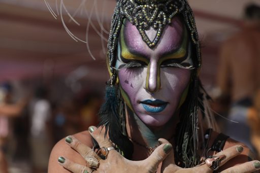 A participant with elaborate face paint and headdress poses with their hands crossed over their chest at Burning Man 2017 in the Black Rock