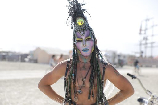 A person with elaborate face paint and a feathered headdress poses with hands on hips, wearing a leather harness and multiple necklaces. The