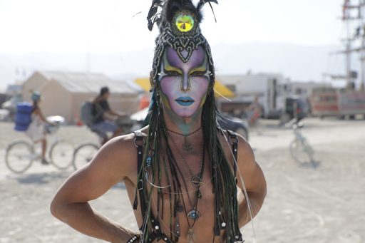 A person with elaborate face paint and headdress poses outdoors at Burning Man 2017. The person wears a black harness and multiple necklaces
