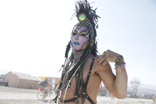 A person with elaborate face paint and a feathered headdress poses outdoors during Burning Man 2017 in Lovelock, Nevada. The individual wear