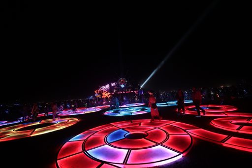 A wide-angle shot captures a nighttime art installation featuring illuminated circular patterns on the ground, with people walking among the