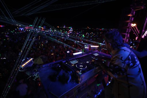 A DJ performs on a stage at night, with a large crowd visible in the background illuminated by numerous laser lights. The DJ is using profes