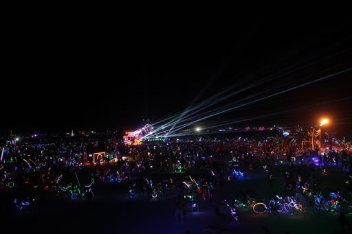 A wide shot captures a nighttime scene at Burning Man 2017 in the Black Rock Desert, Nevada. Thousands of participants, many on illuminated