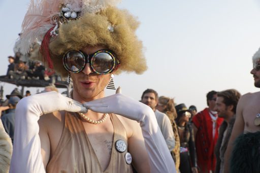 A person in a large, feathered headdress, oversized circular glasses, and white gloves poses with hands near their face at Burning Man in Lo