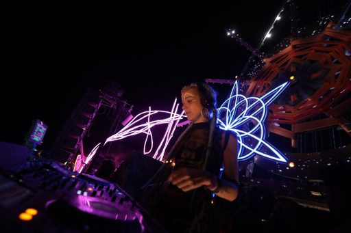 A female DJ performs at night on a stage at Burning Man 2017 in Lovelock, Nevada. She is wearing headphones and standing behind a DJ console