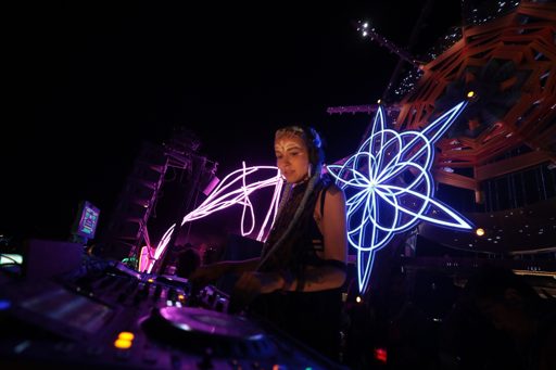 A female DJ performs at night on a stage at Burning Man 2017 in Lovelock, Nevada. She is wearing headphones and has face paint, with a large