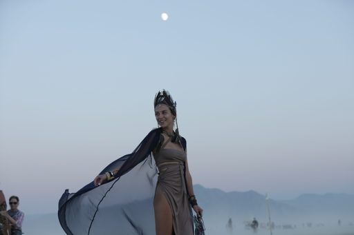 A woman in a crown and flowing cape poses in the desert at dusk, with the moon visible in the pale sky. The scene is part of the Burning Man