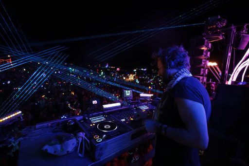 A DJ performs on a stage at night, illuminated by blue laser lights, with a large crowd visible in the background. The scene was captured in