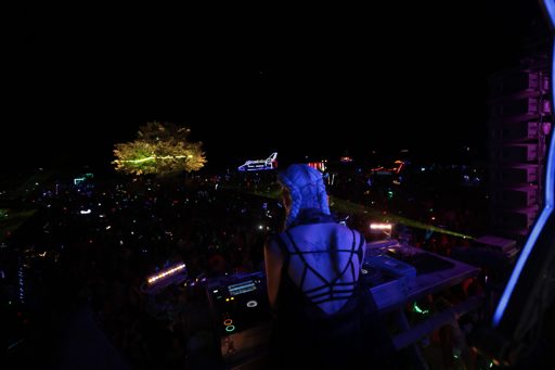 A DJ performs at night in front of a large crowd at Burning Man 2017 in Lovelock, Nevada. A large tree decorated with lights and a structure