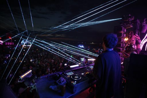 A DJ performs at an outdoor music festival in Lovelock, Nevada, with a large crowd visible in the background and numerous laser beams illumi
