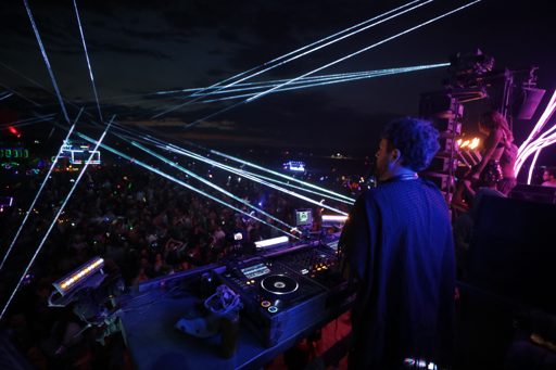 A DJ performs for a large crowd at night, illuminated by numerous white and purple laser beams, during the 2017 Burning Man festival in Love