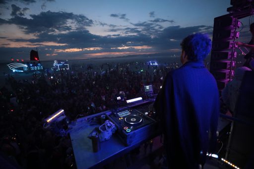 A DJ performs for a large crowd at dusk on the playa, with illuminated art installations in the background. The scene was captured during th