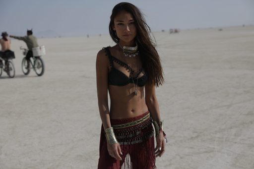 A woman poses in a black bikini top and beaded skirt on the playa during Burning Man 2018. In the background, two people ride bicycles acros