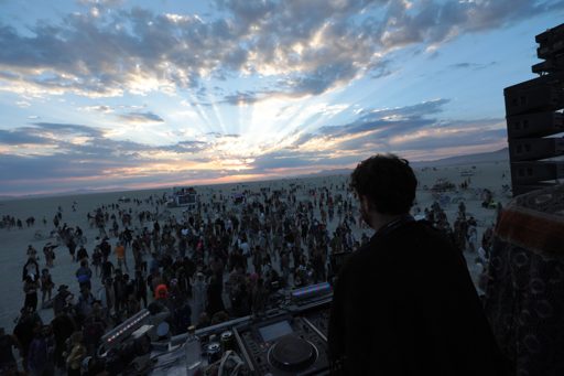 A DJ performs at sunset on the Black Rock Desert playa during the Burning Man festival. The sun's rays beam through dramatic clouds over a v