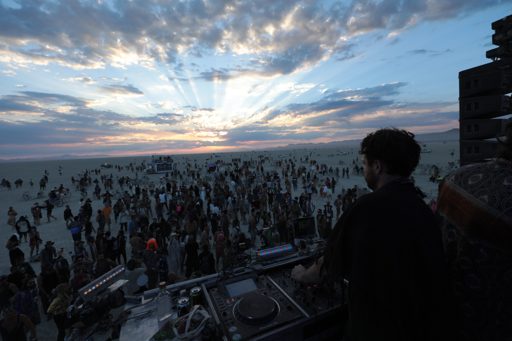 A DJ plays music for a crowd of people gathered on the playa at sunset during Burning Man in Lovelock, Nevada. The sun's rays beam through d