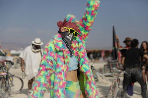 A person in a brightly colored, fuzzy jacket and tie-dye shorts dances with their arm raised at Burning Man 2018. They wear heart-shaped sun