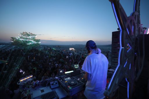 A DJ performs at dusk on a stage at Burning Man 2017, with a large crowd gathered below and a decorated tree illuminated in the distance. Th