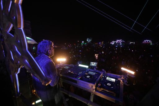 A DJ plays music from a raised platform to a large crowd at night during the Burning Man festival in Lovelock, Nevada. The DJ booth features