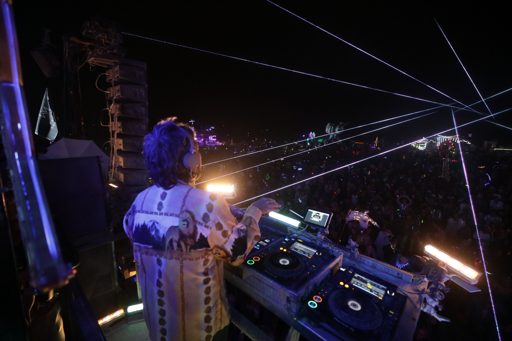 A DJ performs for a large crowd under a night sky filled with laser lights at Burning Man 2017 in Lovelock, Nevada. The DJ, wearing headphon