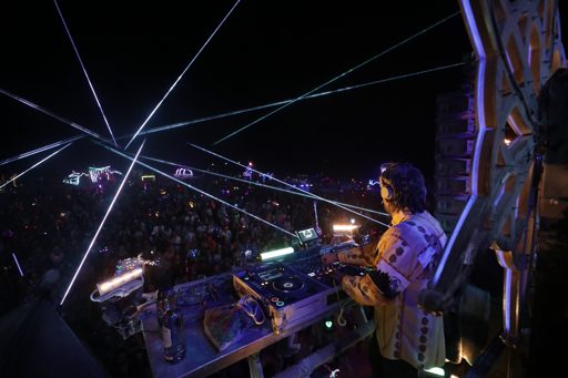 A DJ performs for a large crowd at night, illuminated by numerous white laser beams crisscrossing the sky. The scene is part of the Burning