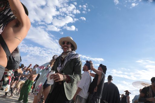A man in a hat and sunglasses laughs while holding a white insulated bottle at an outdoor gathering. The scene is set against a bright blue