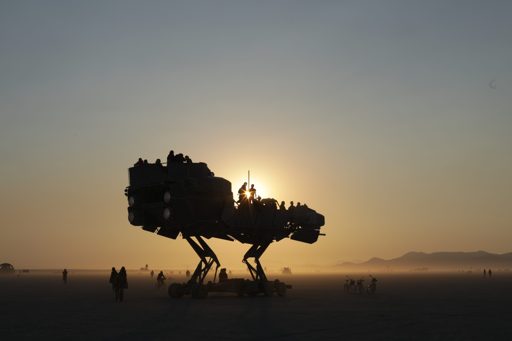 Silhouetted against the setting sun at Burning Man 2017, a large, multi-legged art car, resembling a dinosaur or mechanical beast, carries p