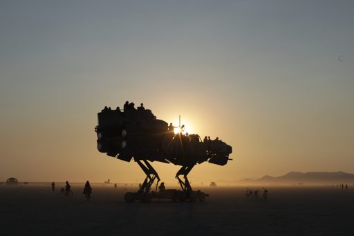 A large, mechanical, dinosaur-like art car, carrying passengers, is silhouetted against the setting sun on the playa during Burning Man 2017