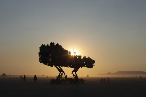 A large, mechanical, dinosaur-like art car, carrying passengers, is silhouetted against the setting sun on the playa during Burning Man 2017