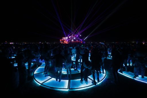 A crowd of people dance on illuminated blue platforms at night, with purple laser lights projecting from a stage in the distance. This scene
