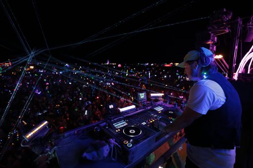 A DJ performs for a large crowd at night, illuminated by numerous laser beams and colorful lights. The DJ is wearing headphones and standing