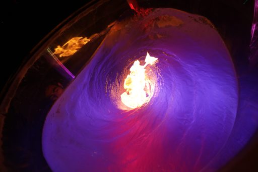 A performer's face is visible in the reflection of a swirling vortex of ice and fire, illuminated by purple and red lights during the Burnin